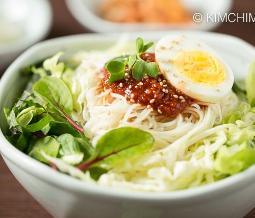 Bowl of bibim guksu served with fresh vegetables , topped with gochujang sauce and egg